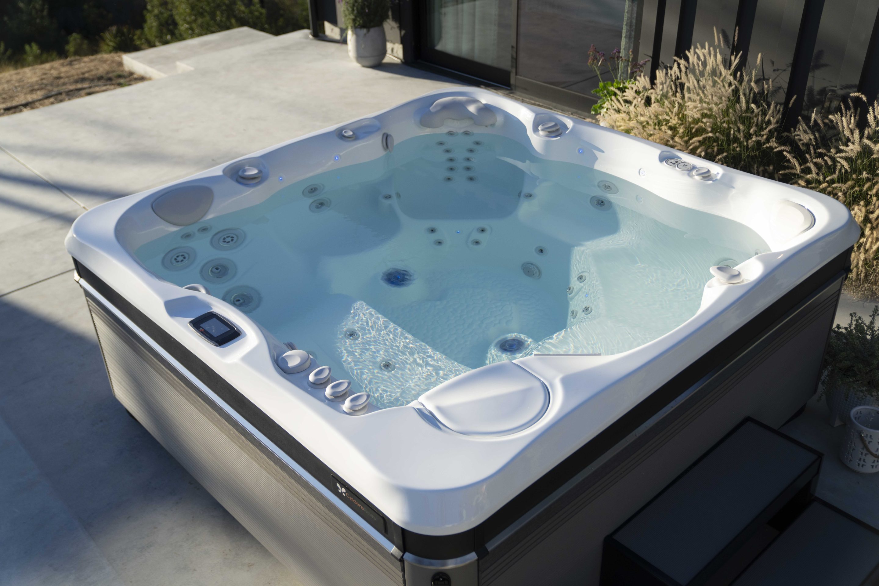 Outdoor white hot tub on a concrete patio beside a modern home, with multiple jets and built-in controls.