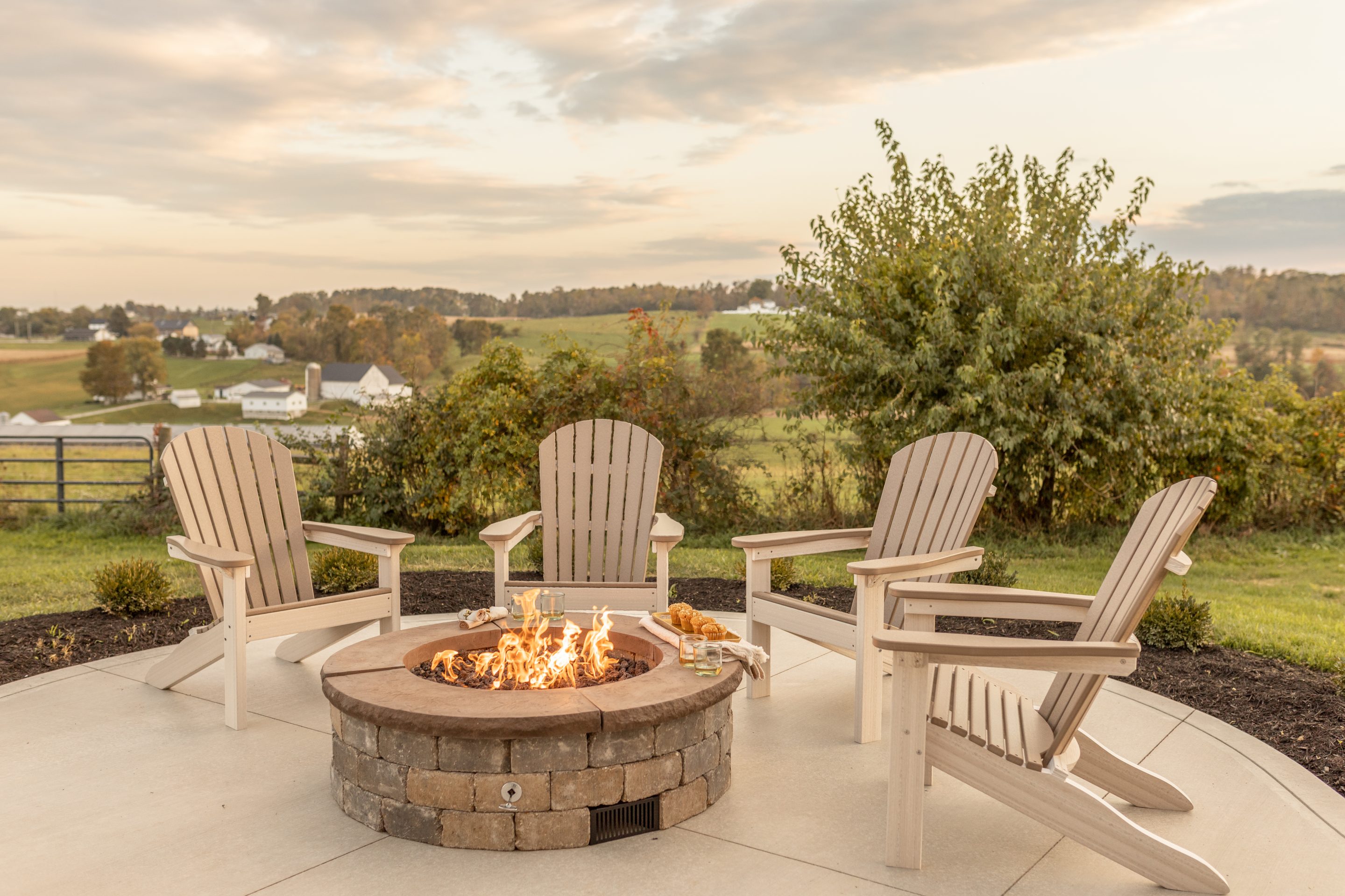 Outdoor patio with a circular stone fire pit surrounded by beige Adirondack chairs overlooking a countryside landscape at sunset.